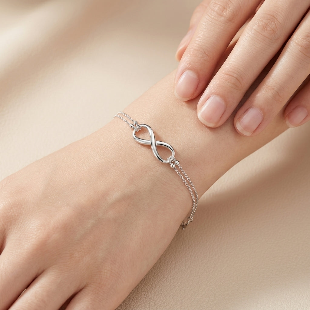 A delicate silver infinity bracelet with a small cluster of clear stones on one side, shown worn on a person's wrist with their fingers resting nearby against a soft beige background.