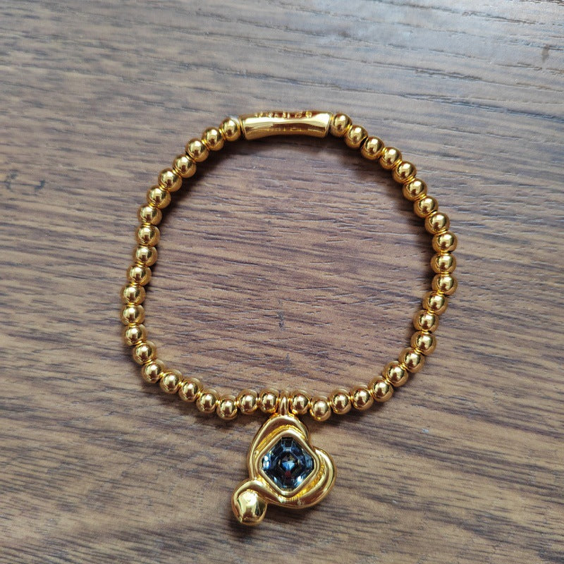 A gold beaded bracelet with a small gold charm featuring a dark blue faceted gemstone, displayed on a brown wooden surface.