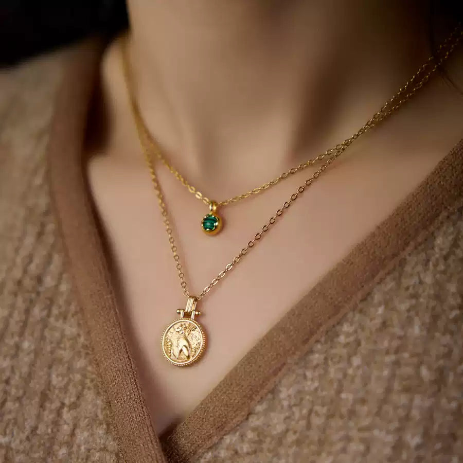 Two gold necklaces with a green gemstone and a coin pendant on a neutral background