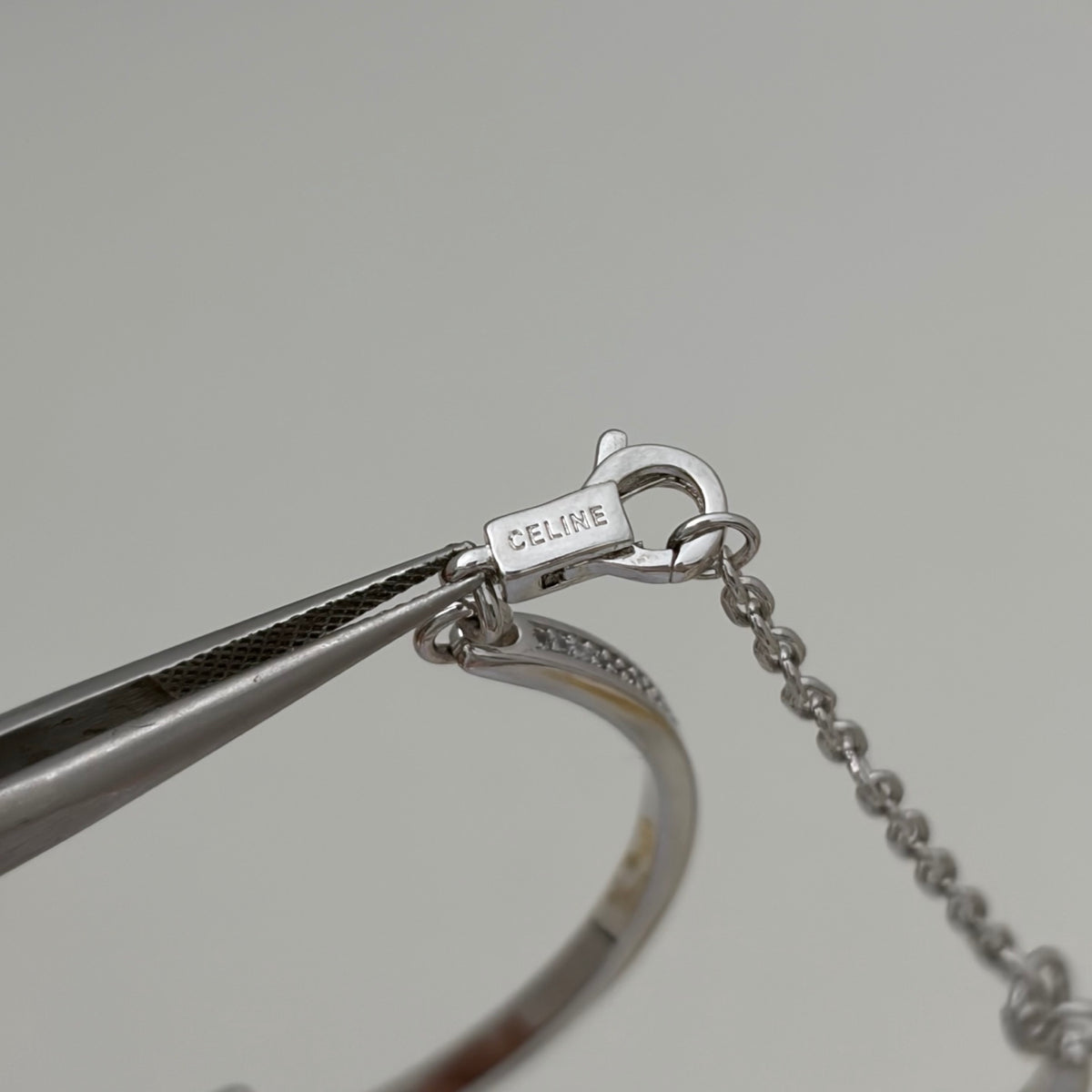 Close-up of a silver bangle bracelet with a lobster clasp engraved with 'CELINE', held by metal tweezers against a light gray background.