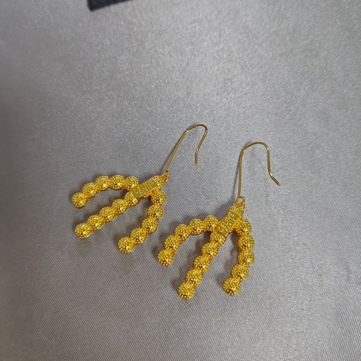A pair of gold-tone dangle earrings with textured, beaded branch-like drops, displayed on a neutral gray textured fabric background.