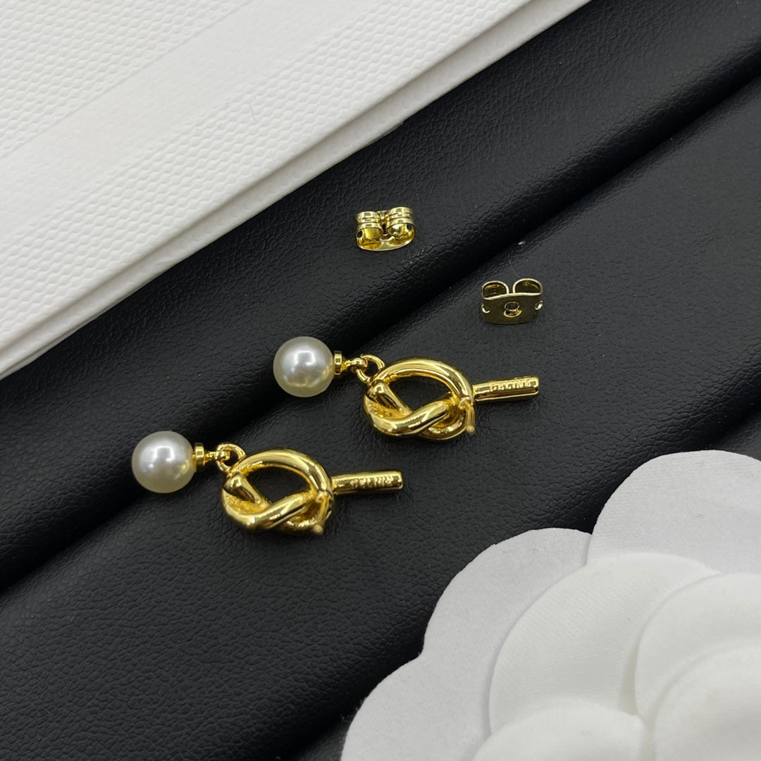 A pair of gold-toned toggle stud earrings with white pearl drops, displayed on a black textured surface with white paper and flower petals as styling props.