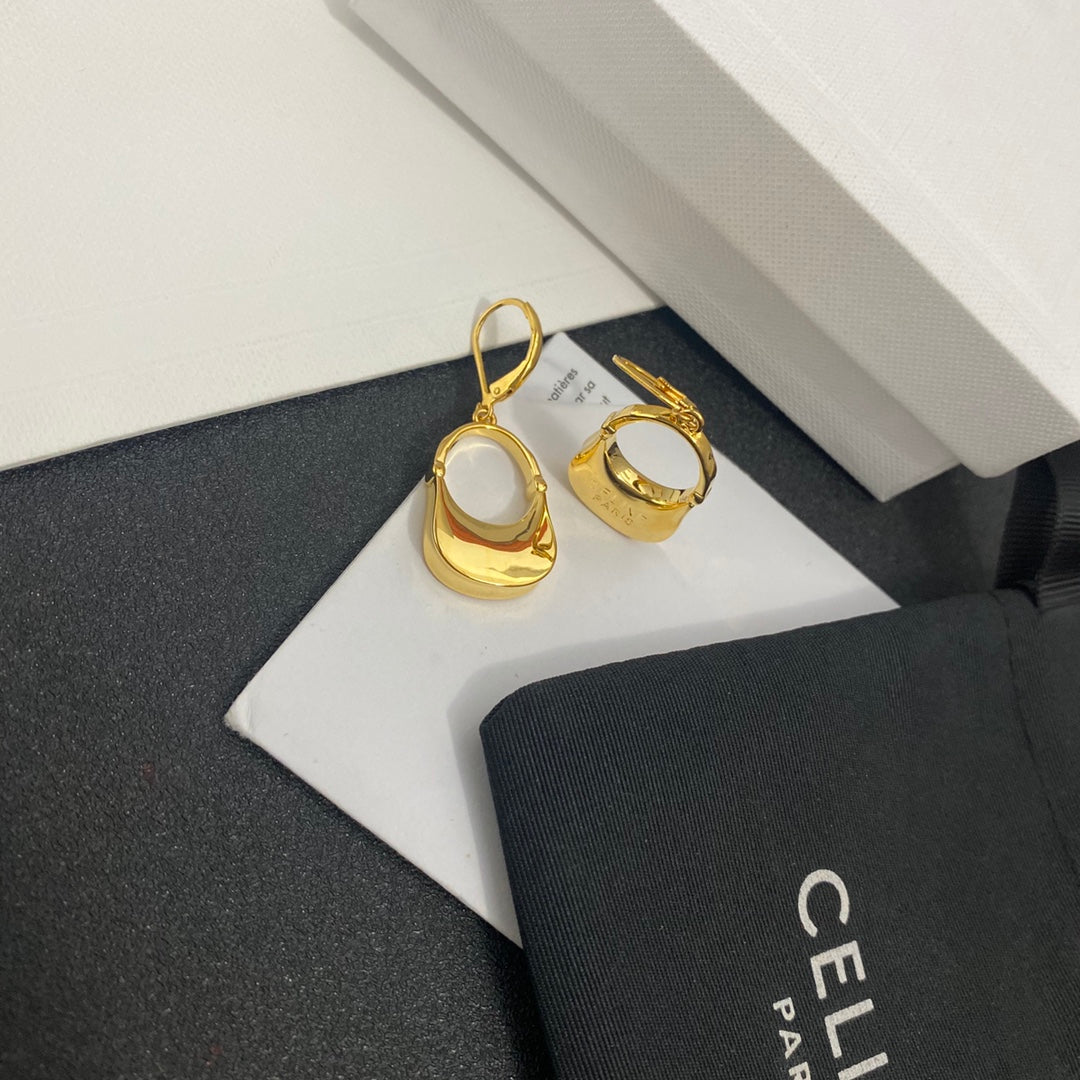 A pair of gold crescent-shaped hoop earrings displayed on a white card, with a black CELINE Paris dust bag and a white textured box in the background.