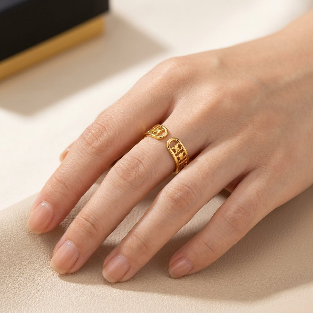 A close-up of a hand wearing a gold ring with a textured, openwork geometric design, resting on a light beige textured surface.