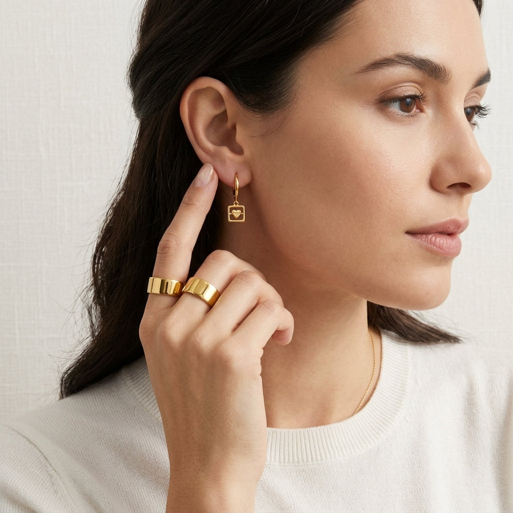 A close-up lifestyle shot of a woman wearing a delicate gold drop earring with a square charm featuring a monogram logo, along with two wide gold rings on her fingers. She is wearing a cream-colored knit top against a textured off-white background.