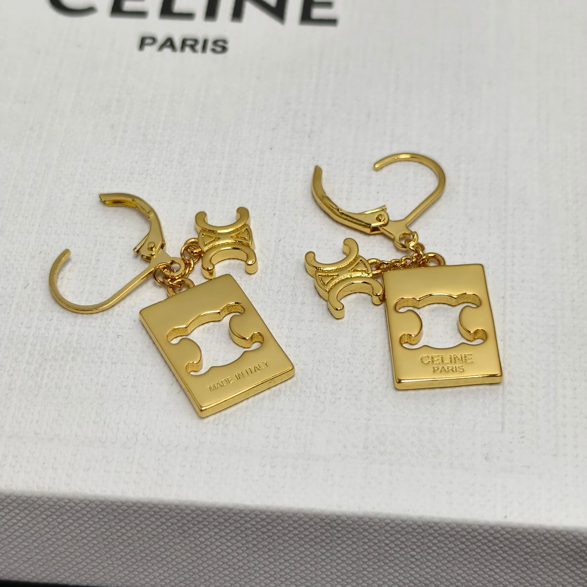 A pair of gold-tone Celine drop earrings with rectangular logo charms and leverback hooks, displayed on a white textured Celine box.