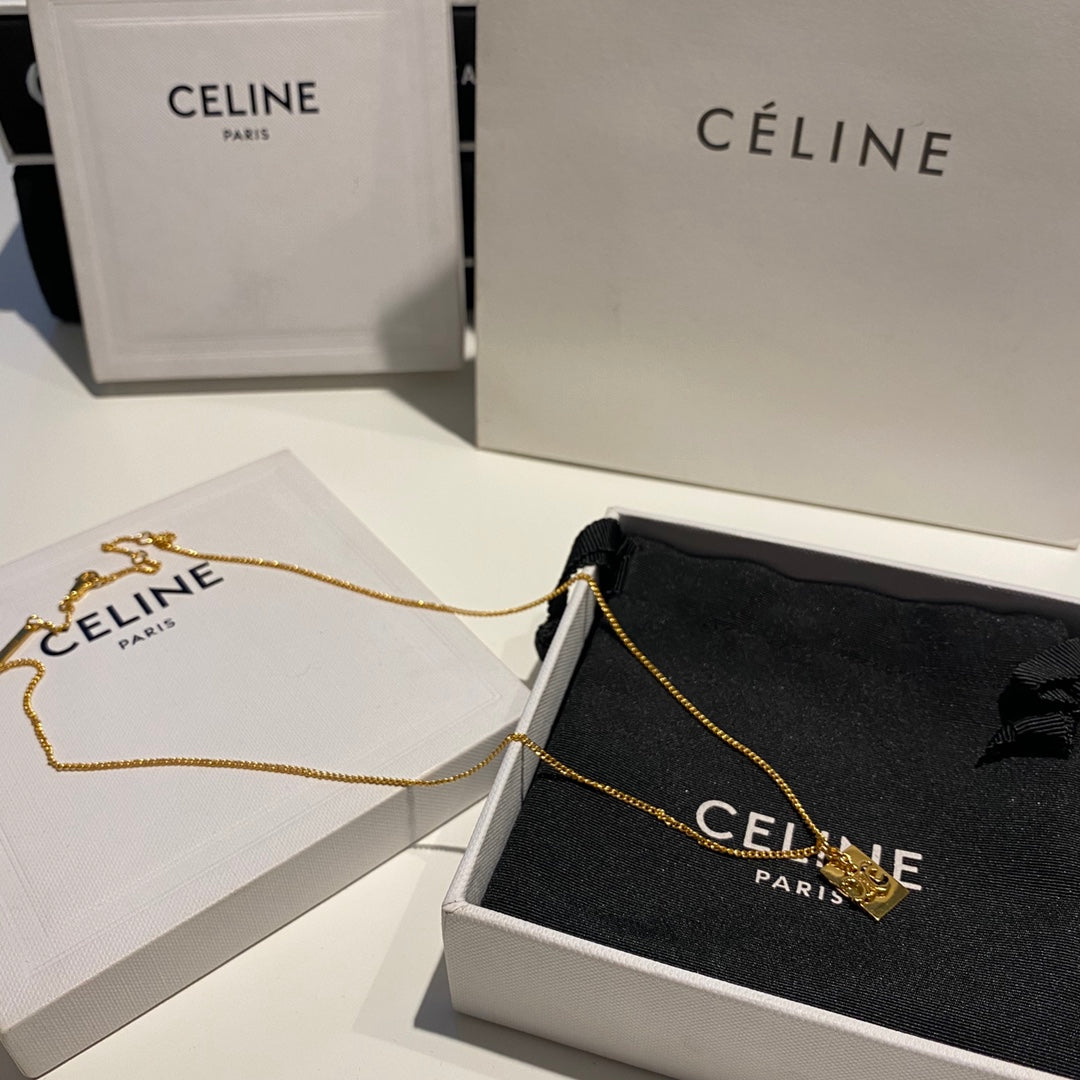 A gold-tone Céline necklace with a small heart-shaped charm, displayed on a black Céline dust bag inside an open white Céline box, with additional Céline packaging in the background.