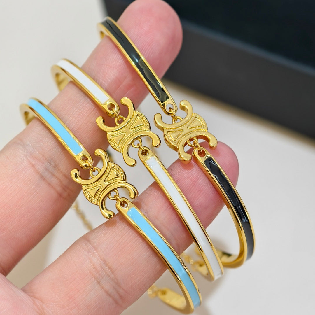 Three gold-plated bangle bracelets with white, light blue, and black enamel stripes, each featuring a decorative gold charm, held on a person's fingers.
