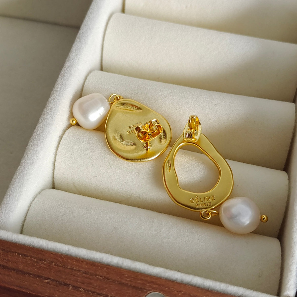 A pair of gold dangle earrings with irregular white freshwater pearls, displayed inside a cream-colored jewelry box.