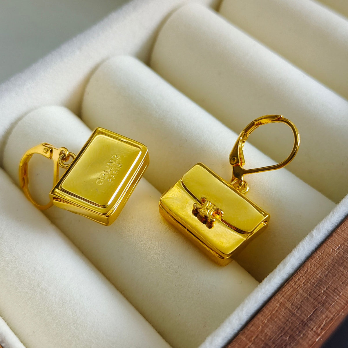 A pair of gold rectangular box-shaped earrings displayed in a cream-colored jewelry box. One earring is shown from the front with a small engraved brand mark, and the other is shown from the side with its hook clasp.