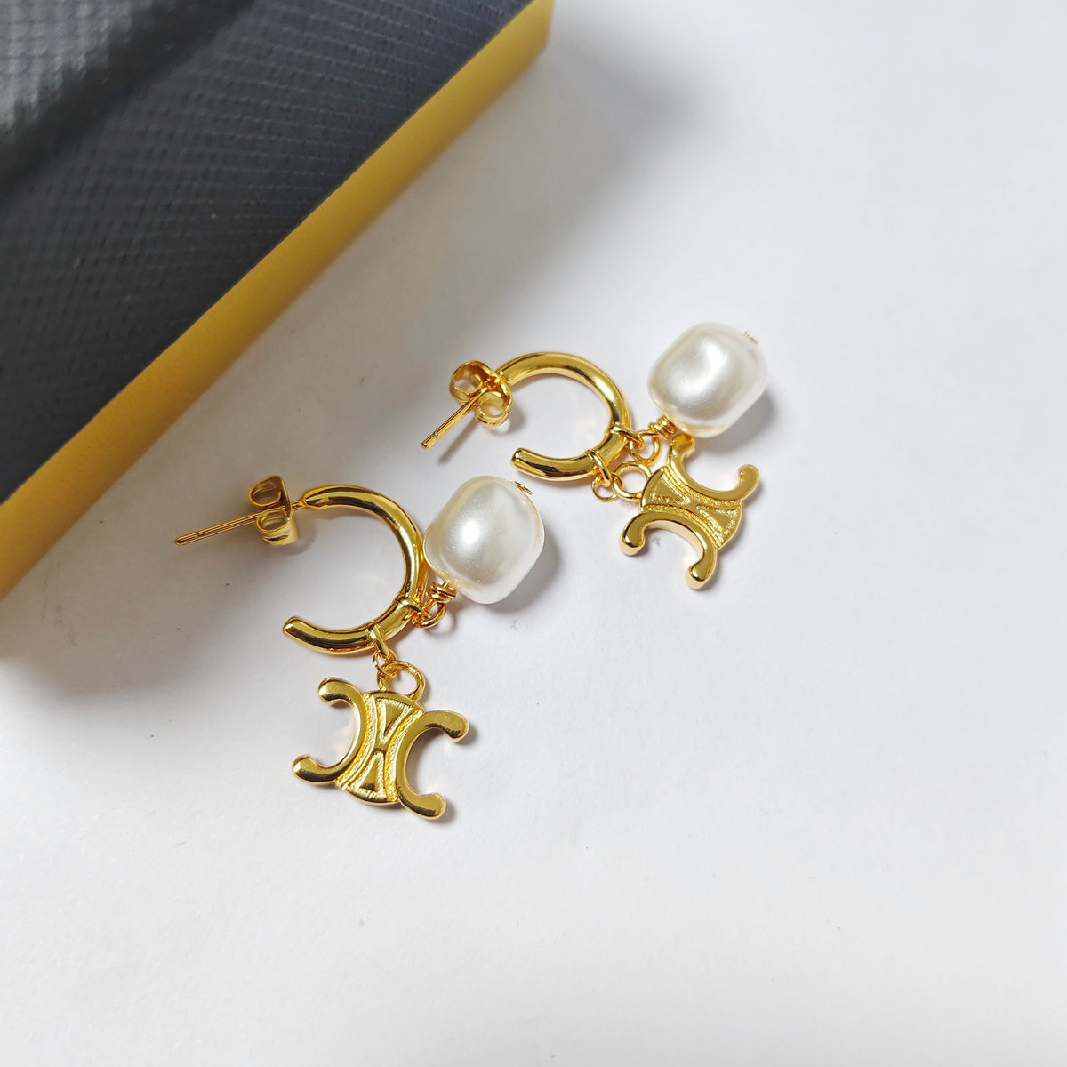 A pair of gold-tone hoop earrings with a dangling Celine Triomphe emblem and a white freshwater pearl, displayed on a white surface next to a yellow and black box.