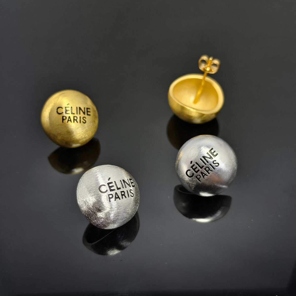 Three spherical Céline Paris stud earrings displayed on a glossy black reflective surface, including one gold-toned earring with its backing visible and two silver-toned earrings.