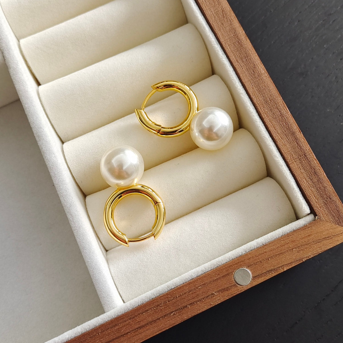 A pair of gold huggie hoop earrings with white freshwater pearls, displayed on a cream velvet-lined wooden jewelry box.