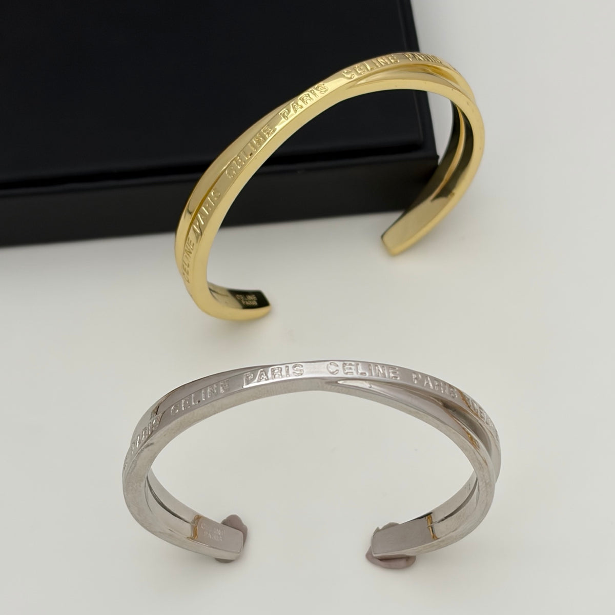 Two open cuff bracelets displayed on a white surface, one in polished yellow gold and one in polished silver, both engraved with 'PARIS CELARE'. A black jewelry box is partially visible in the background.