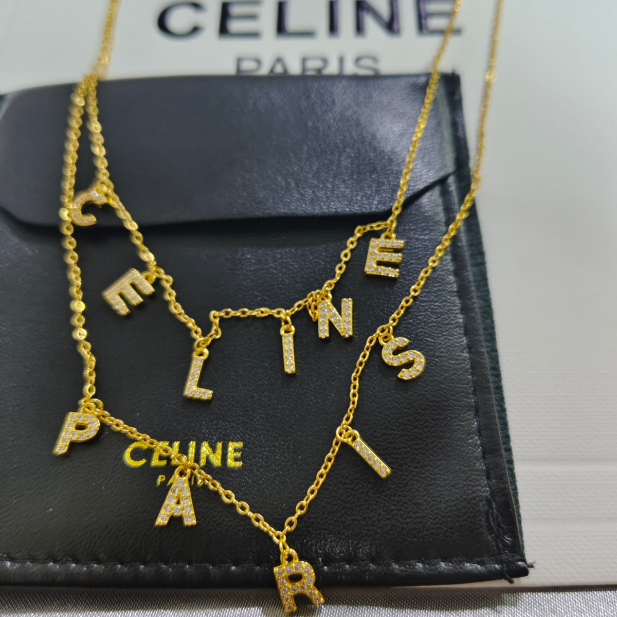 A gold-tone Celine letter necklace with pavé-set stones, draped over a black Celine pouch with the brand name visible.