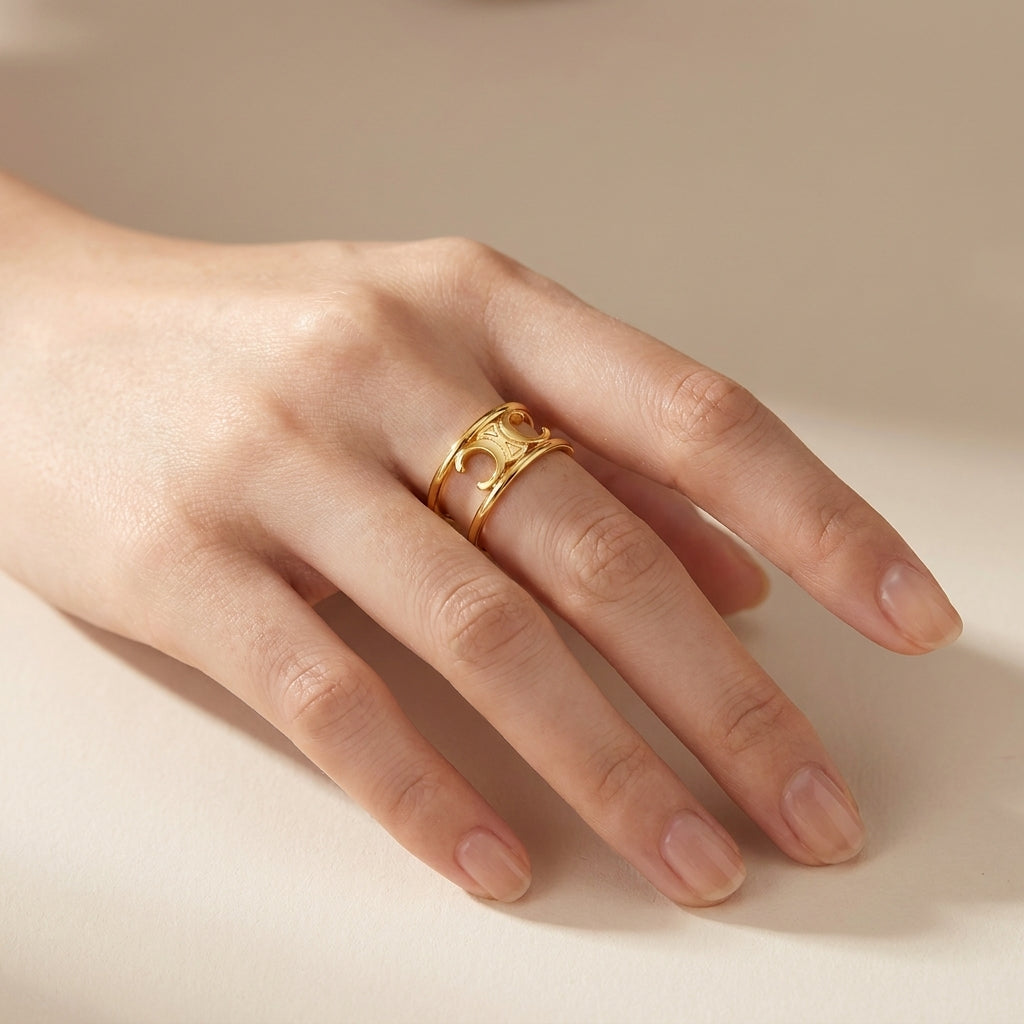 A close-up of a hand wearing a gold ring with a textured, organic openwork design, set against a soft beige background.