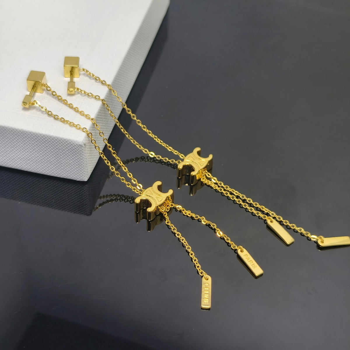 A pair of gold-toned long dangle earrings displayed on a dark reflective surface next to a white display block. Each earring features a square stud, a delicate chain, a small pagoda charm, and two rectangular dangling tags.