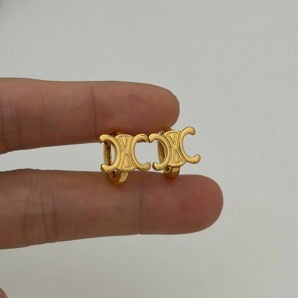 A pair of gold-tone Celine double-C logo hoop earrings held between two fingers against a plain light gray background.