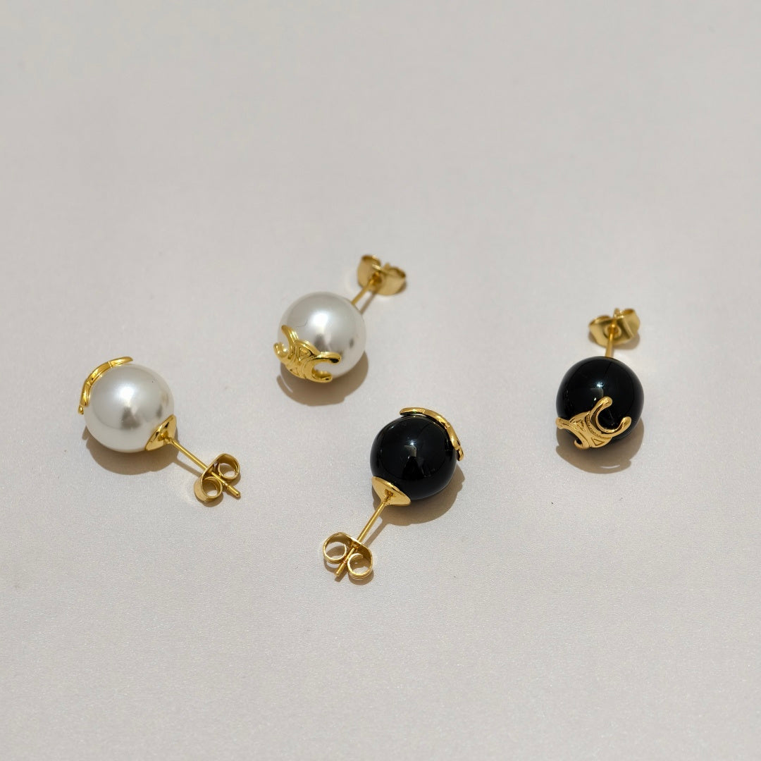 Four gold-tone stud earrings arranged on a light gray background, including two white pearl studs and two glossy black bead studs.