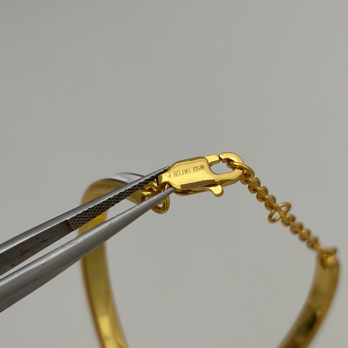 A close-up of a gold bangle bracelet with an extension chain and lobster clasp, held by silver tweezers against a neutral light gray background. The inside of the clasp is stamped with 'MADE IN INDIA'.