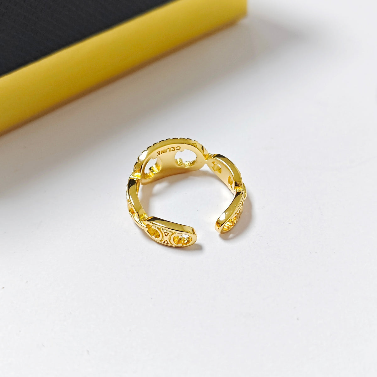 A gold-toned Celine open ring with a textured, ribbed design and the CELINE brand name engraved inside, resting on a white surface with a yellow and black geometric prop in the background.
