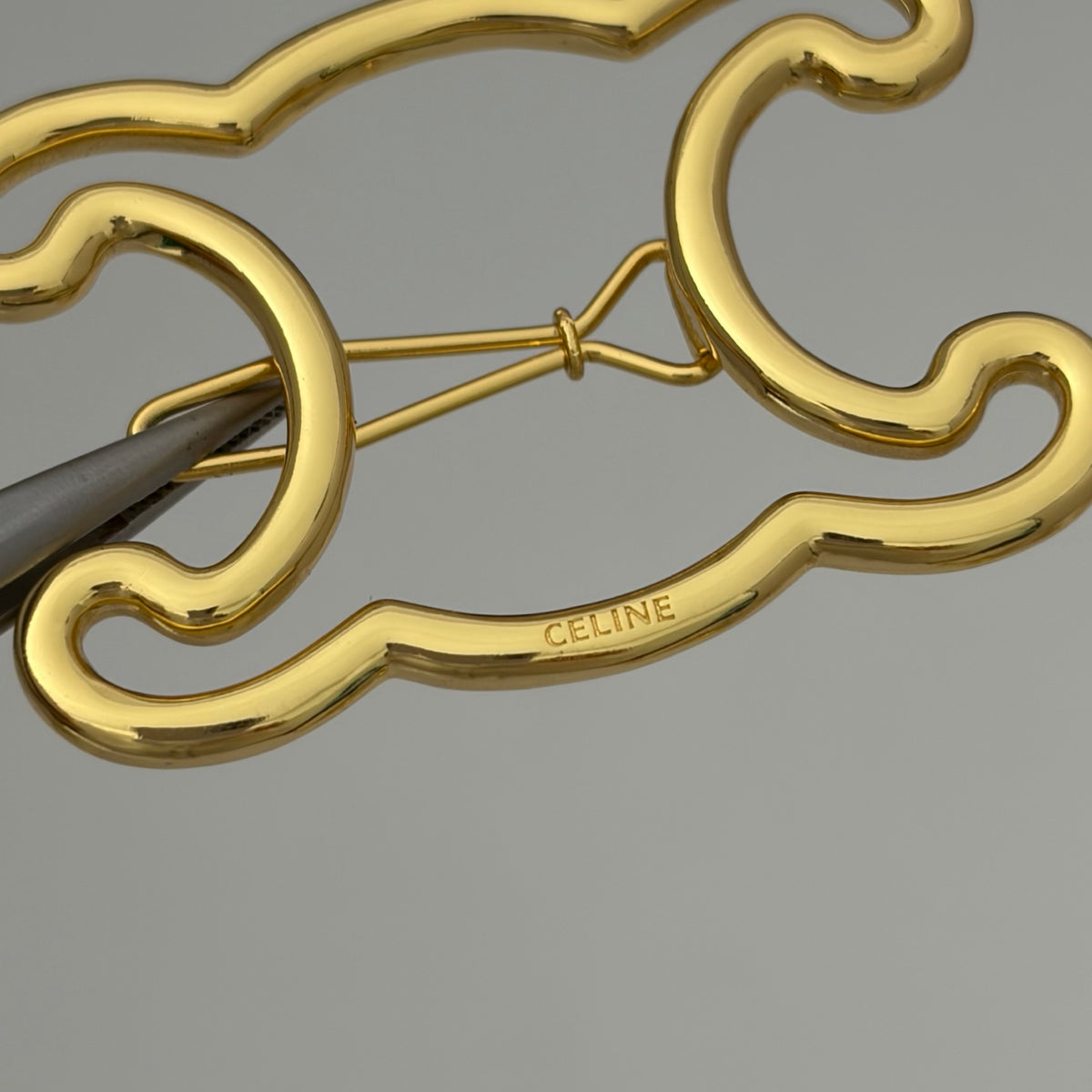 A close-up of a gold-tone Celine logo pin with tweezers holding it in place, showing the engraved CELINE brand name on a neutral gray background.