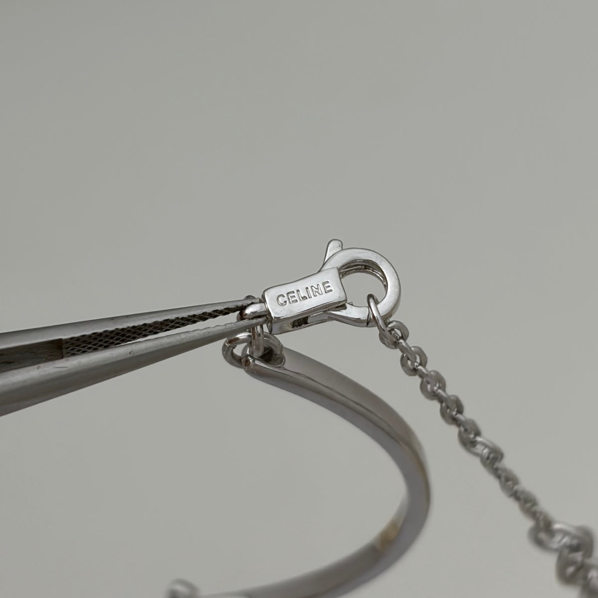Close-up of a silver-toned bangle bracelet with a lobster clasp engraved with 'CELINE', held by tweezers against a neutral gray background.