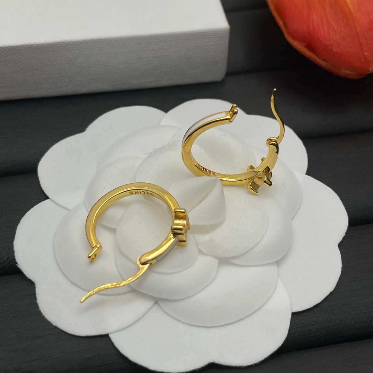 A pair of gold hoop earrings displayed on a white flower-shaped stand, with a white box and a red flower petal partially visible in the background.