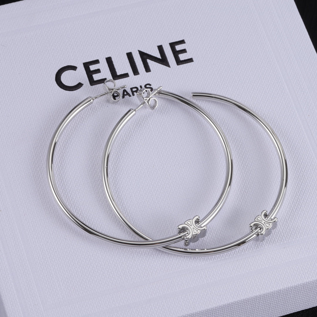 A pair of silver-tone hoop earrings with small fleur-de-lis charms, displayed on a white textured surface printed with the Celine logo.