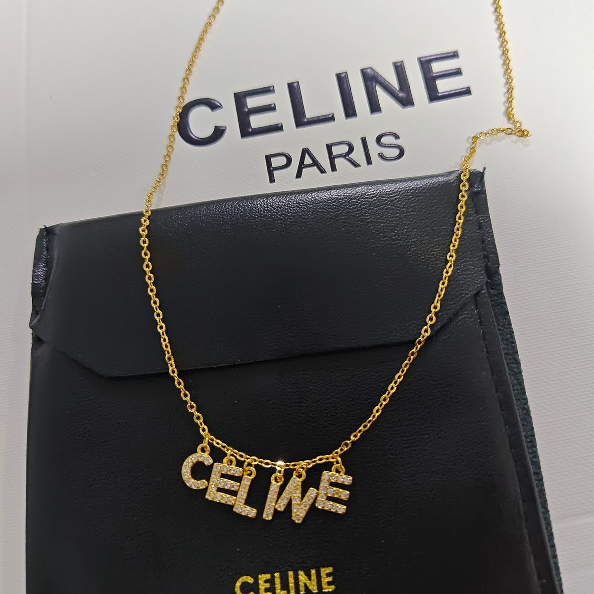 A gold-tone Celine name necklace spelling 'CELINE' in sparkling letter charms, draped over a black Celine pouch with the Celine Paris logo visible in the background.