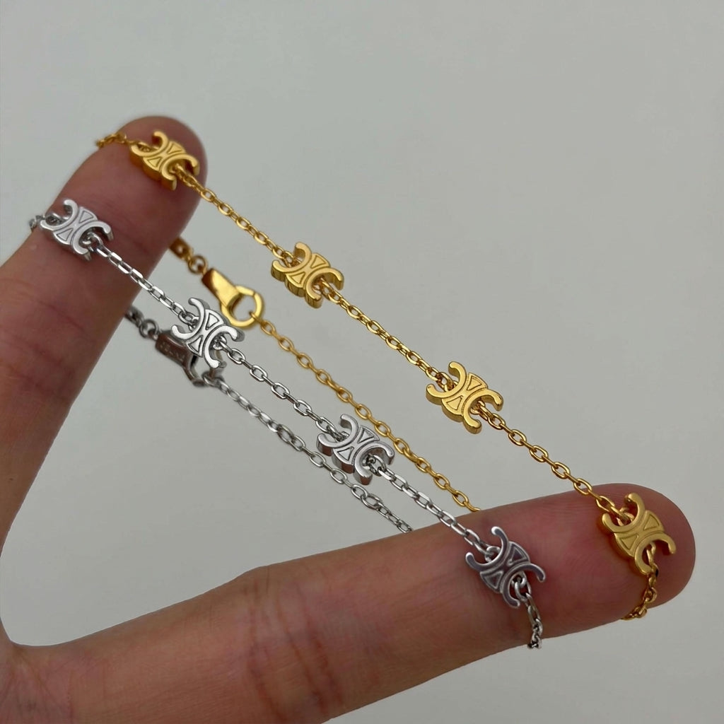 A close-up of a hand holding two delicate chain bracelets, one gold and one silver, both featuring ornate fleur-de-lis motifs.
