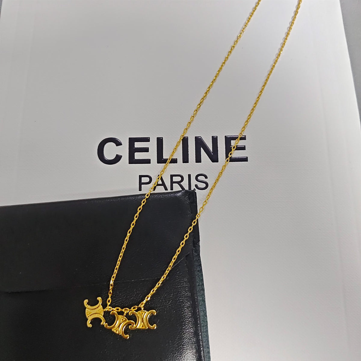A gold-tone Celine necklace with a signature triple-C logo pendant, draped over a black leather box with a white Celine Paris box in the background.