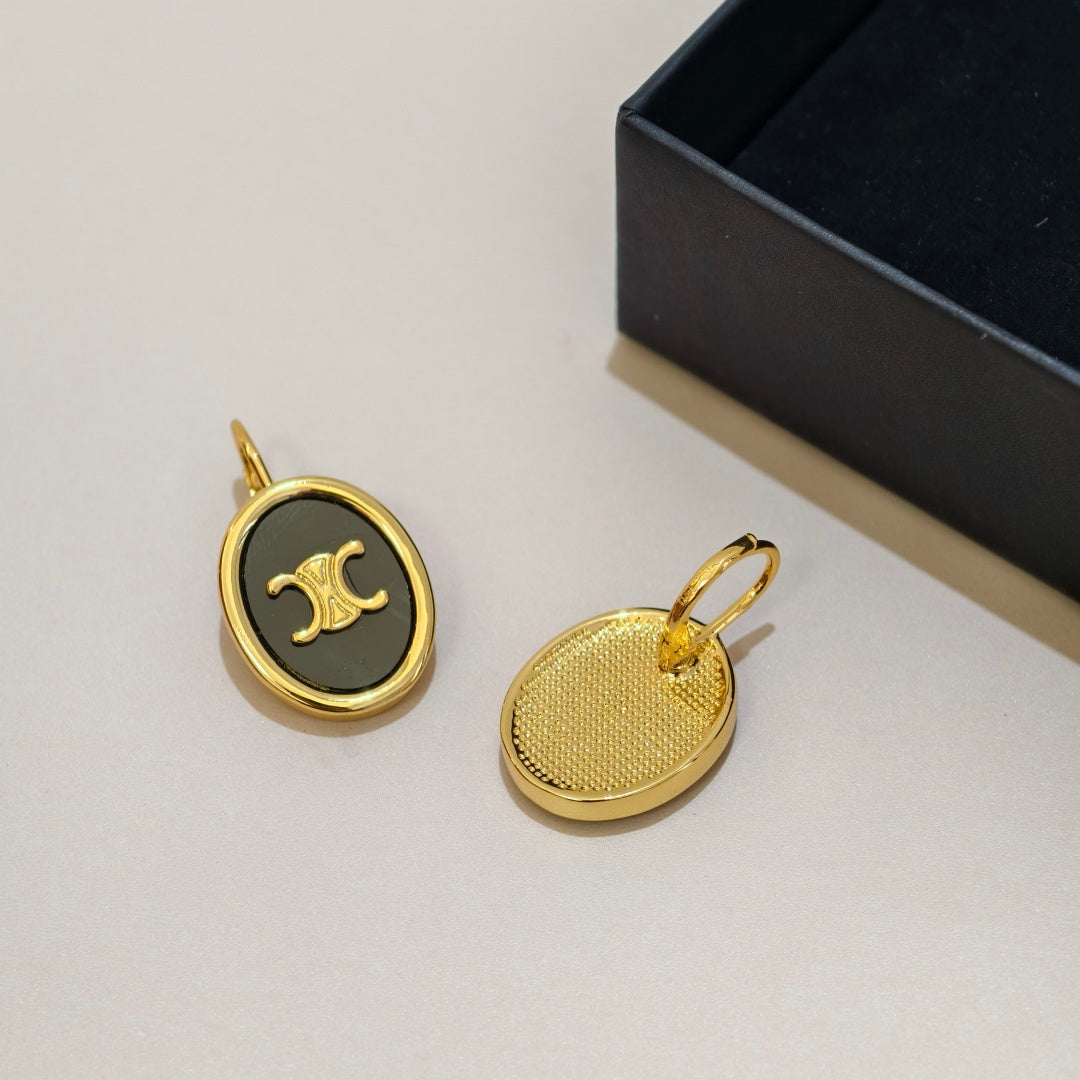 Two oval gold-tone earrings with black enamel inserts and a gold logo emblem, displayed next to a black presentation box on a light beige surface.