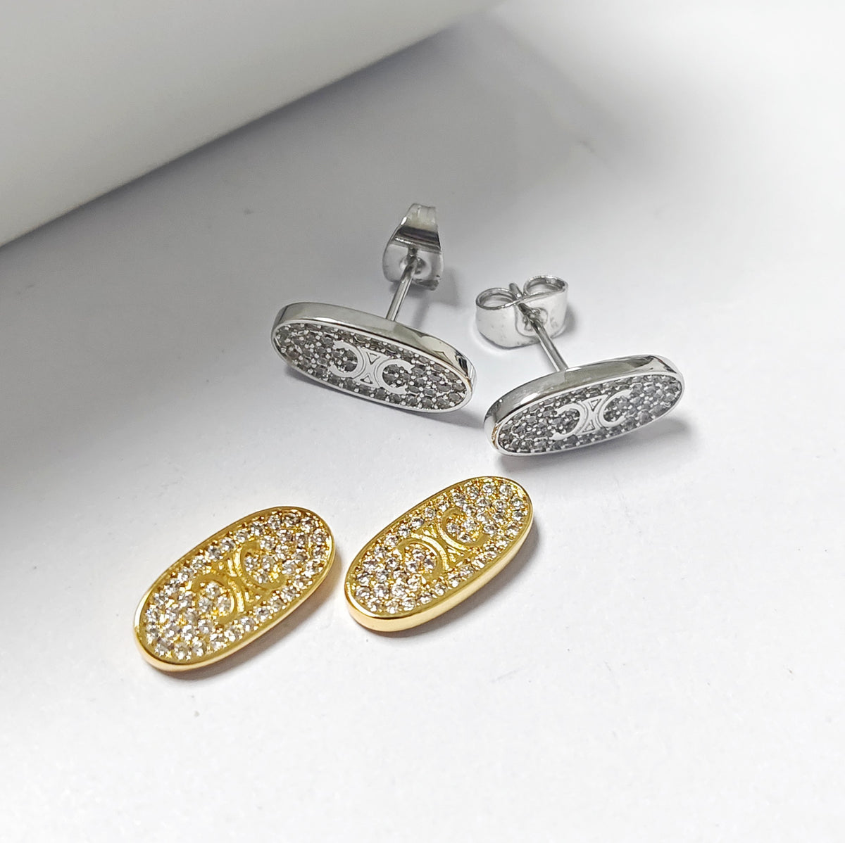 Two pairs of oval stud earrings on a clean white surface. One pair is yellow gold with pavé-set clear stones, and the other is silver-toned with matching pavé detailing.