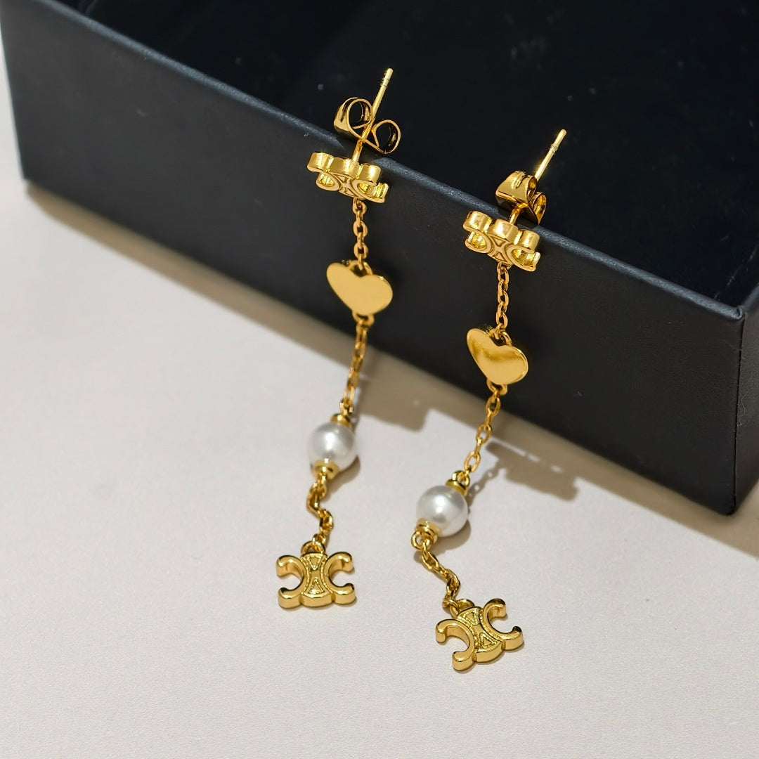 A pair of gold dangle earrings with heart charms, white pearl beads, and small gold charms, displayed on a light surface next to a black box.