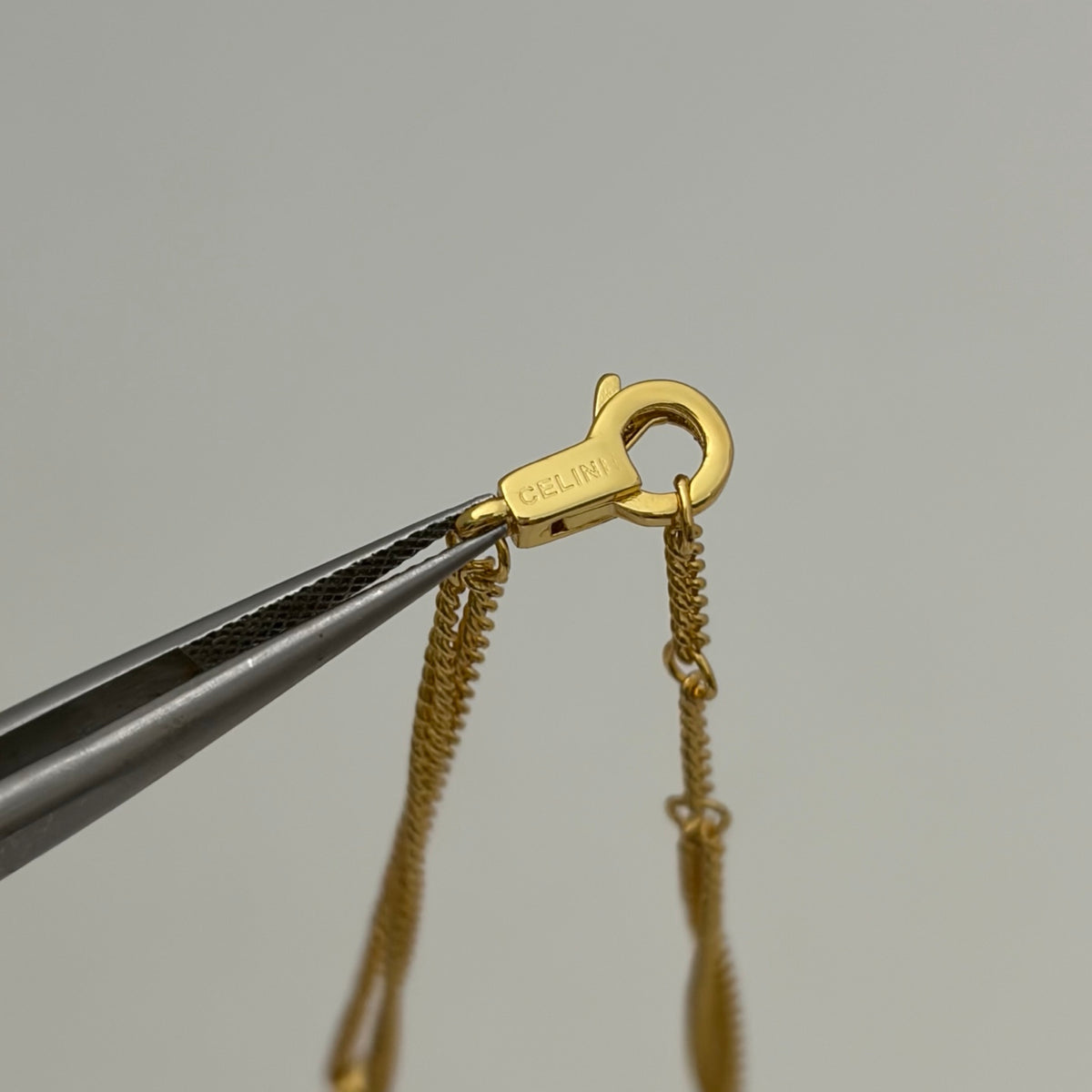 A close-up of a gold lobster clasp held by metal tweezers against a plain light gray background. The clasp is engraved with the brand name CELINI.