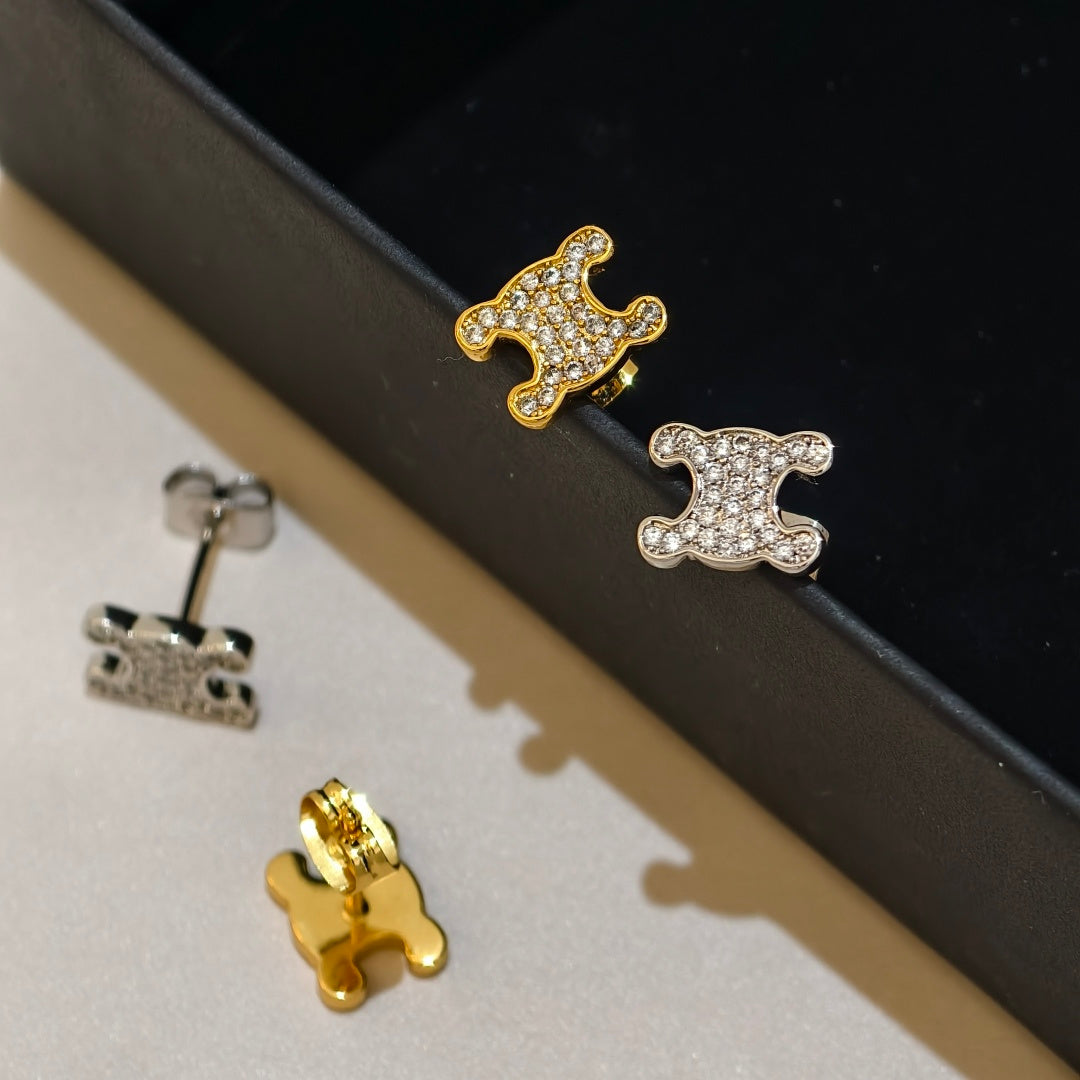 Three small stud earrings shaped like abstract animal heads or faces, shown on a black display tray and beige surface. Two earrings are gold-toned with clear stones, and one is silver-toned with clear stones; the backings are also visible.