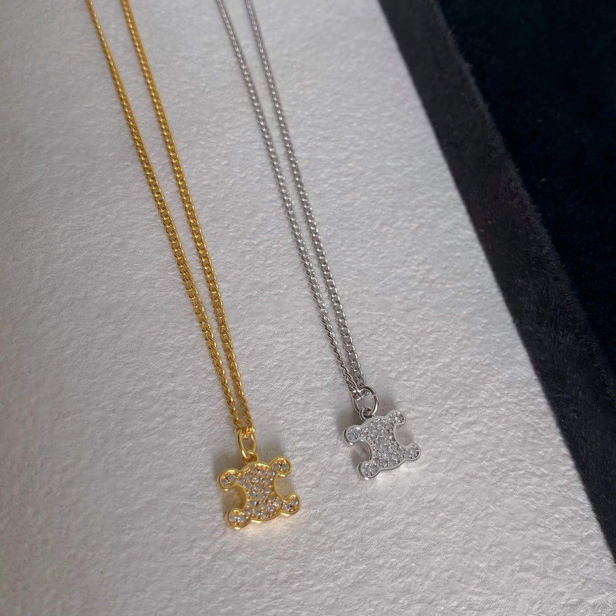 Two fashion necklaces with a matching interlocking CC logo pendant, one in yellow gold and one in silver, both paved with small clear stones, displayed on a textured white surface.