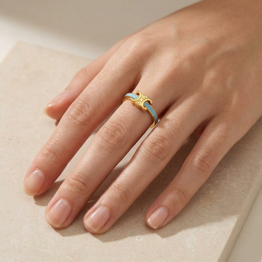 A close-up of a hand wearing a delicate gold ring with a light blue inlay, resting on a neutral beige stone surface.