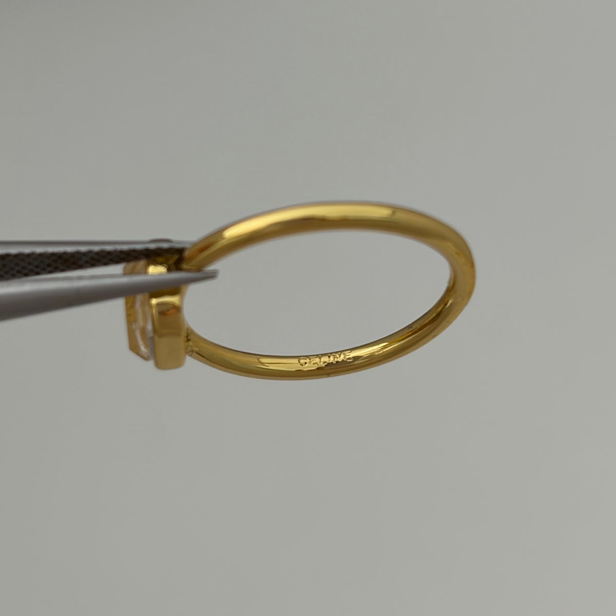 A close-up of a polished yellow gold ring held by metal tweezers against a plain light gray background. The inside of the band features a small, faint engraved hallmark.