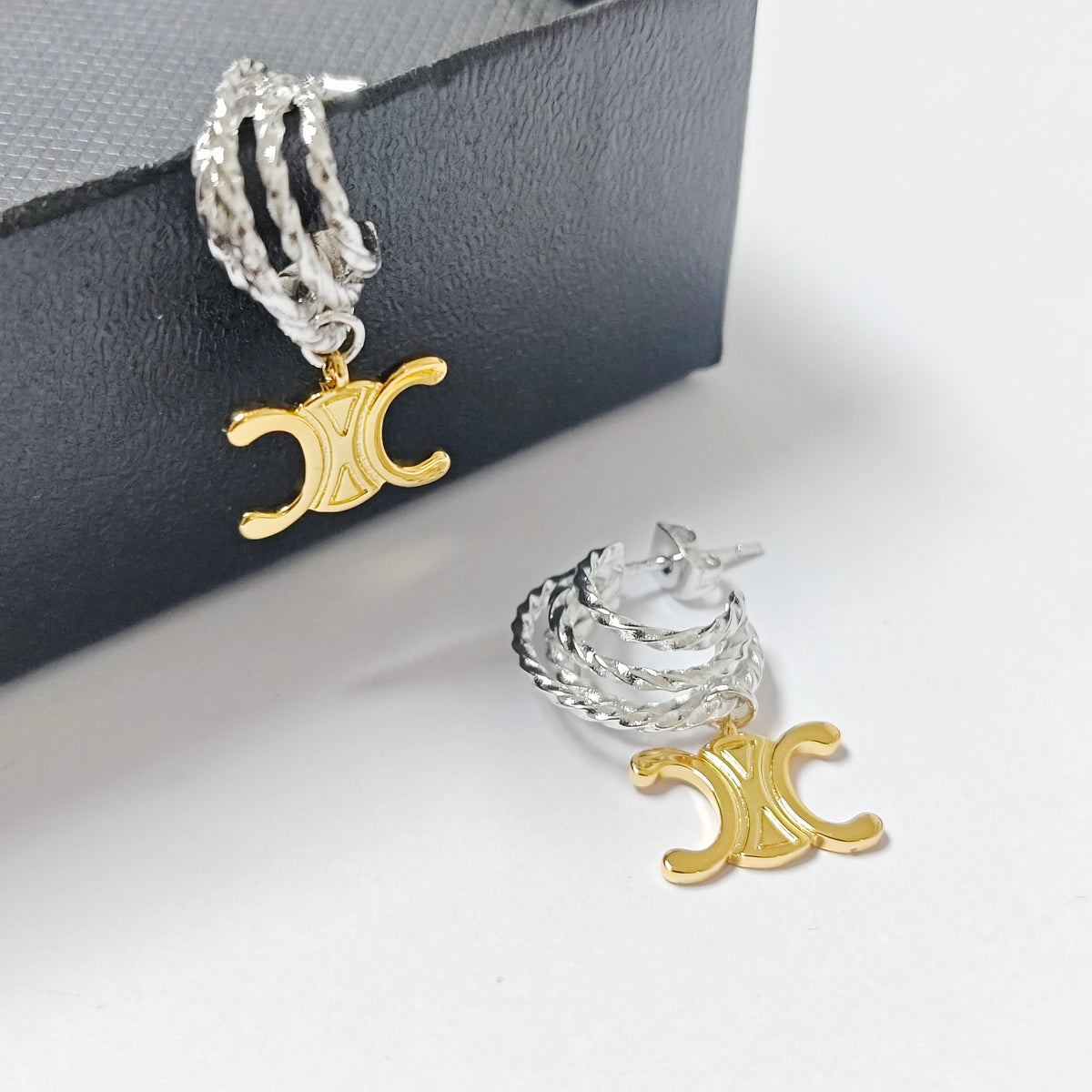 A pair of silver and gold triple-layered hoop earrings with a gold triple-C logo charm, displayed on a white surface with a black textured box in the background.