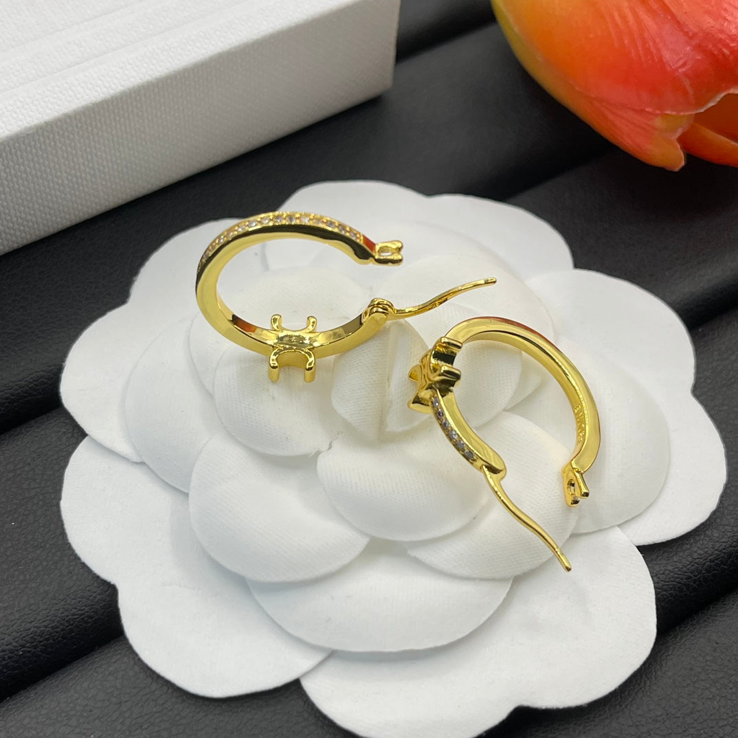 A pair of gold-toned hoop earrings with small clear stones, displayed on a white fabric flower against a dark textured background.