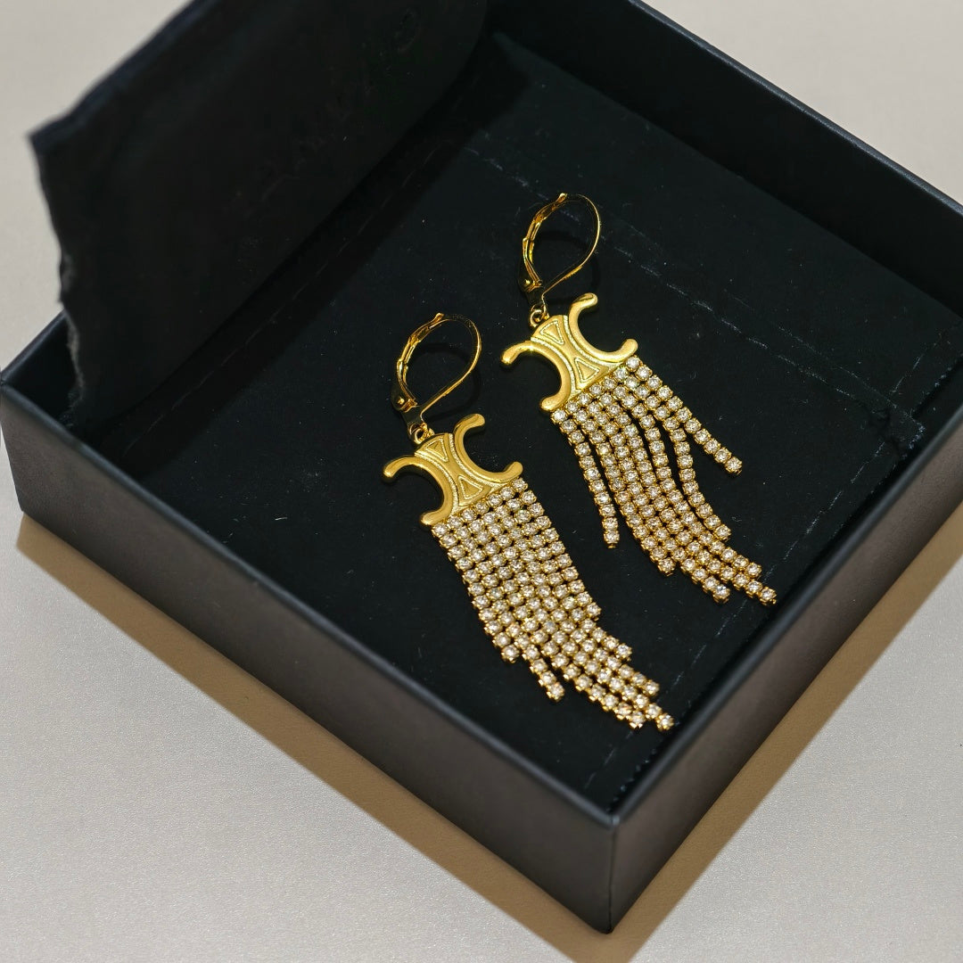 A pair of gold drop earrings with intricate beaded fringe, displayed inside an open black presentation box on a light beige surface.