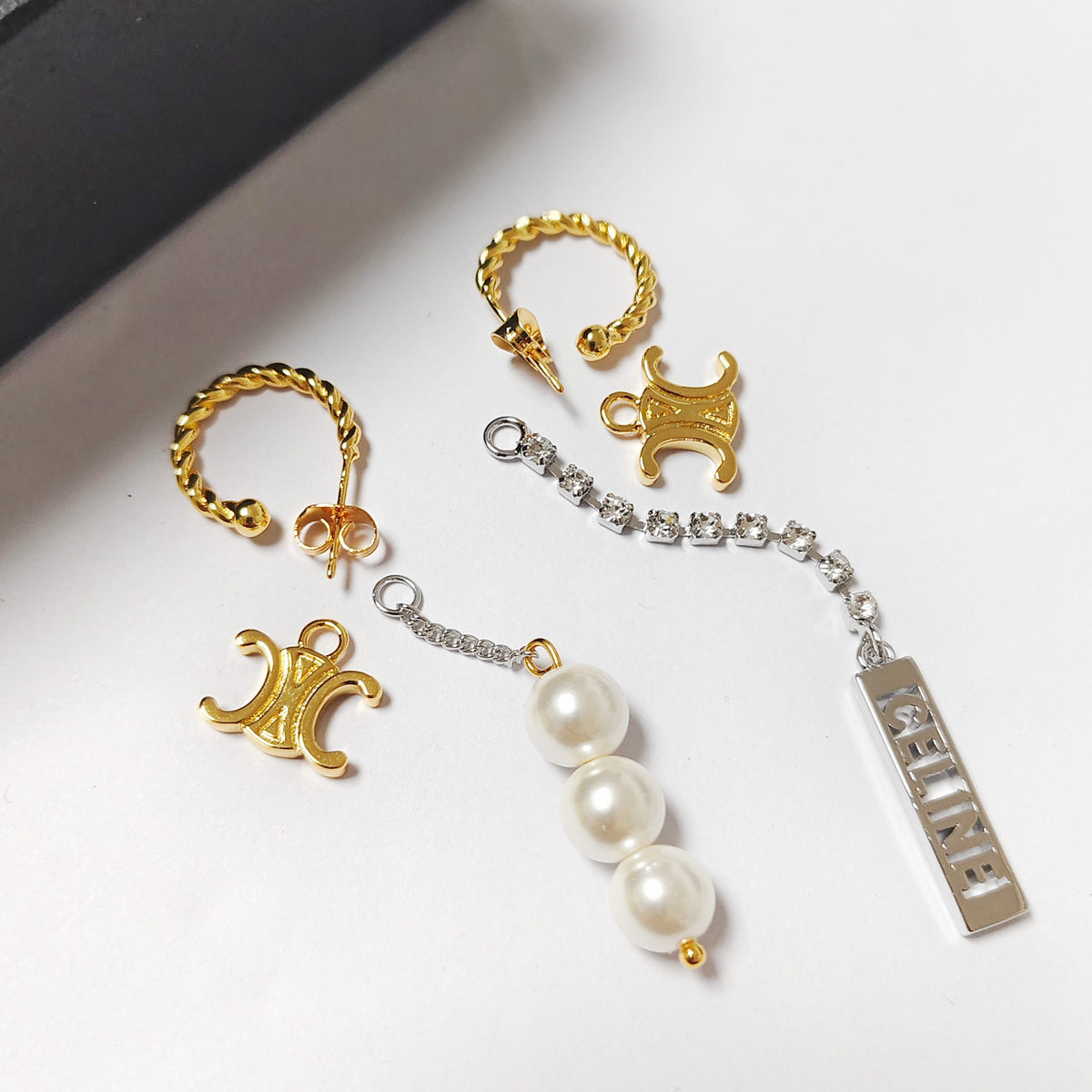 A pair of gold hoop earrings with a matching gold trident charm, displayed alongside a silver chain earring featuring three white faux pearls and a rectangular silver nameplate engraved with 'HELINE'.