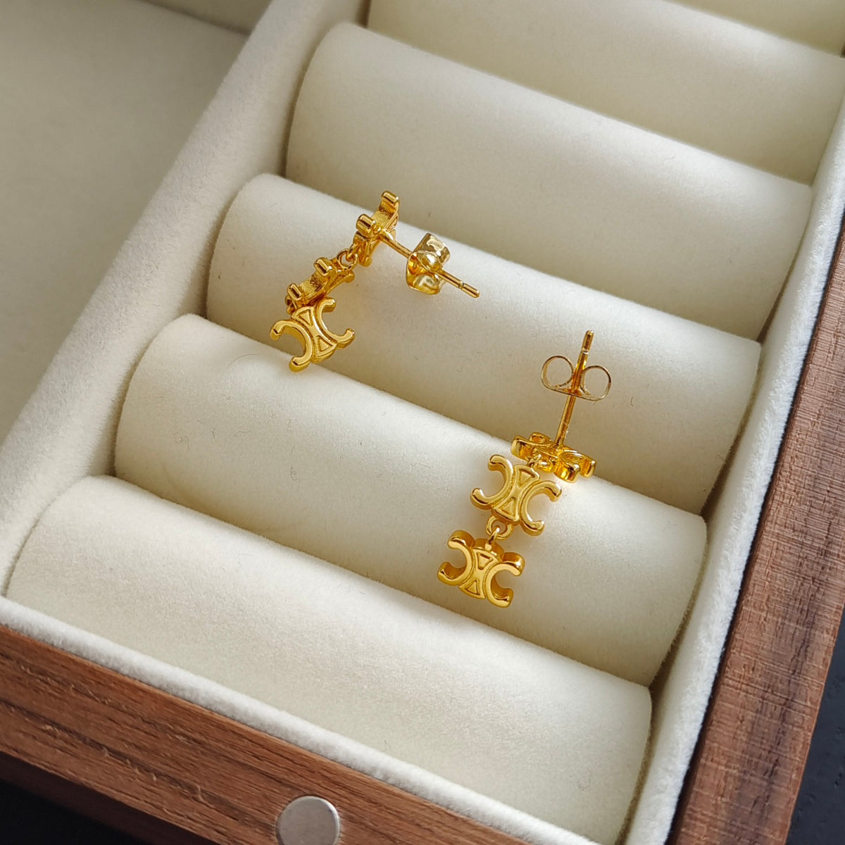A pair of gold drop earrings with ornate C-shaped charms, displayed on cream-colored cylindrical cushions inside a wooden jewelry box.