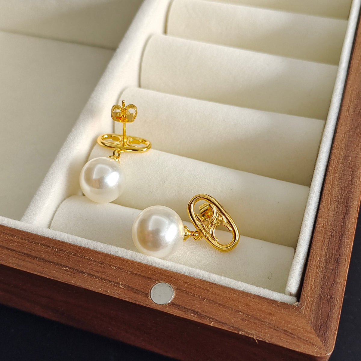 A pair of gold hoop earrings with white freshwater pearls, displayed inside a wooden jewelry box with cream velvet compartments.