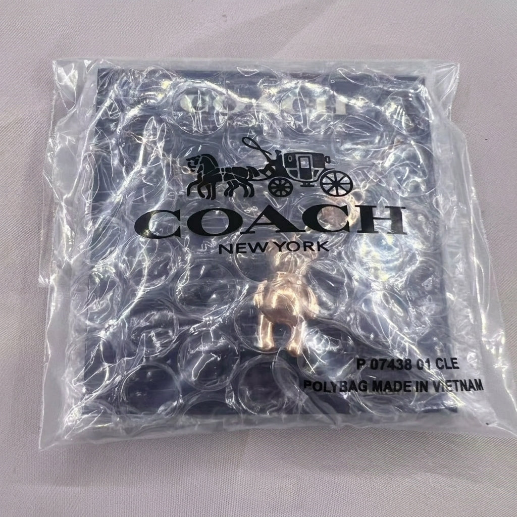 A Coach New York branded clear plastic poly bag containing a gold-tone horse logo charm, photographed on a light fabric background.