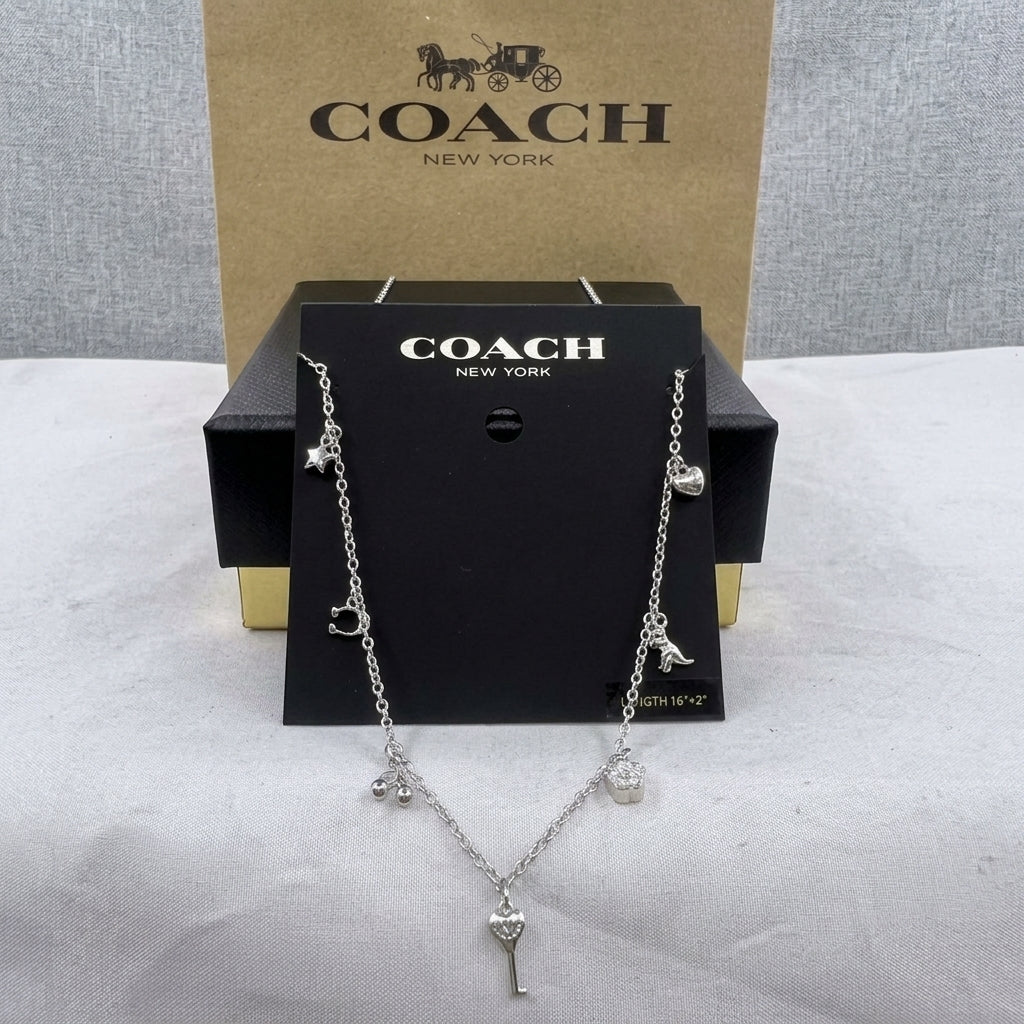 A silver-tone Coach necklace with charms including a carriage, star, bird, and key, displayed on a black Coach card with the Coach New York box behind it.