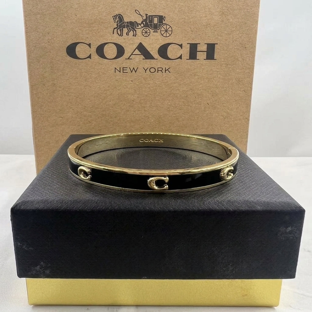 A Coach bangle bracelet with a gold-tone finish and black enamel sections, displayed on a black presentation box with a brown Coach New York box behind it.