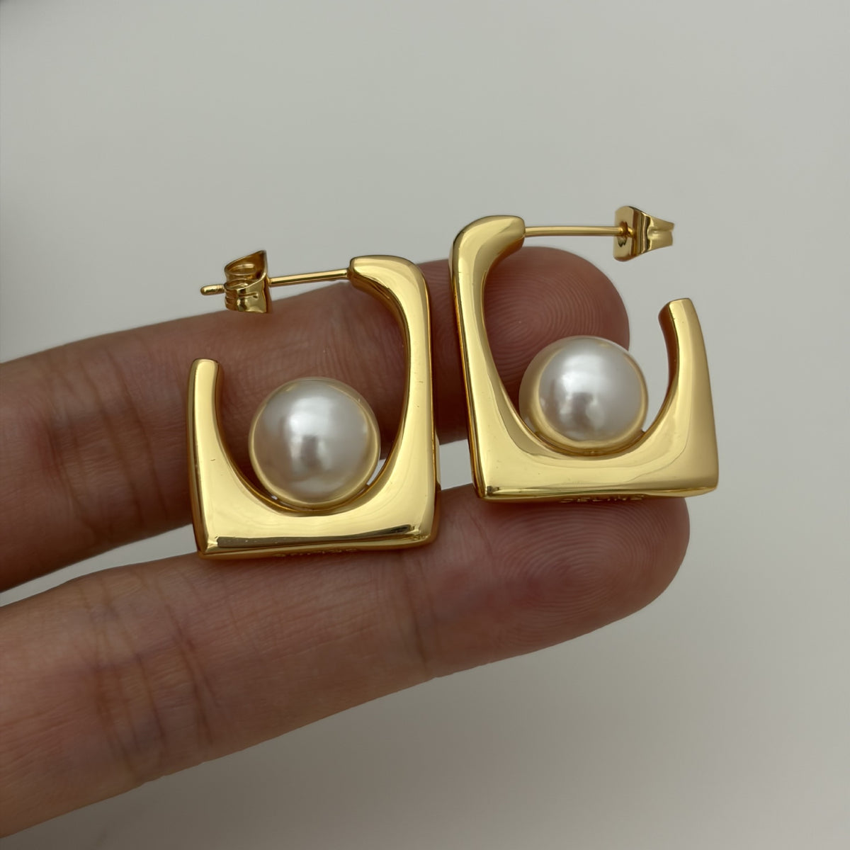 A pair of gold hoop-style earrings with white pearl centers, shown resting on a person's fingers against a plain light background.